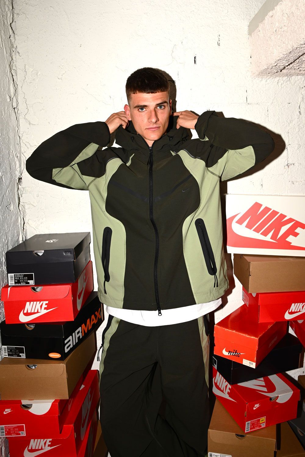 Alex Quijada, vestido con un chándal verde y negro, está de pie en el interior, entre pilas de cajas de zapatillas Nike, ajustándose la capucha con ambas manos y mirando a la cámara. Le rodean paredes blancas con textura.