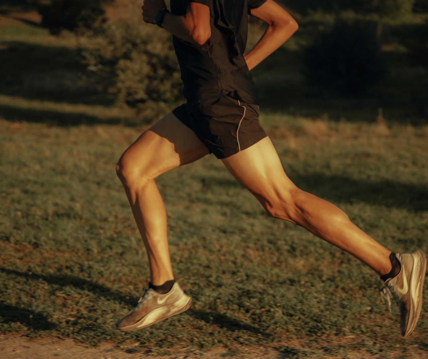 Alex Quijada está corriendo al aire libre por un sendero cubierto de hierba, vestido con unos pantalones cortos negros y una camiseta oscura; solo se le ven las piernas y parte del torso a la cálida luz del día.