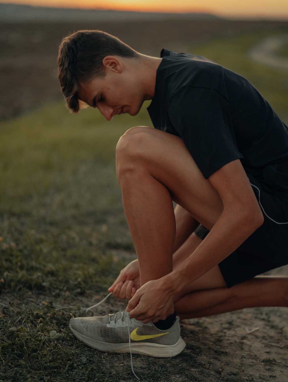Alex Quijada, vestido con camiseta y pantalones cortos negros, está arrodillado en un camino de tierra al atardecer, atándose las zapatillas de correr grises, con la hierba y las colinas extendiéndose al fondo.