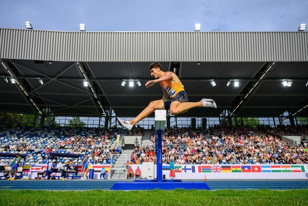 Alex Quijada, un atleta vestido con una camiseta amarilla y pantalones cortos azules, salta una valla de carrera de obstáculos en una pista azul, en un estadio repleto de espectadores, con banderas nacionales ondeando a lo largo de las gradas.