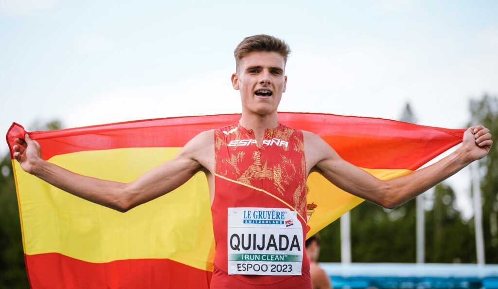 Un deportista, Alex Quijada, vestido con un uniforme rojo y amarillo, sostiene la bandera española que hay detrás de él. Está sonriendo y en su dorsal se lee «Quijada, ESPAÑA, ESPoo 2023». Al fondo se ven árboles y un cielo azul.