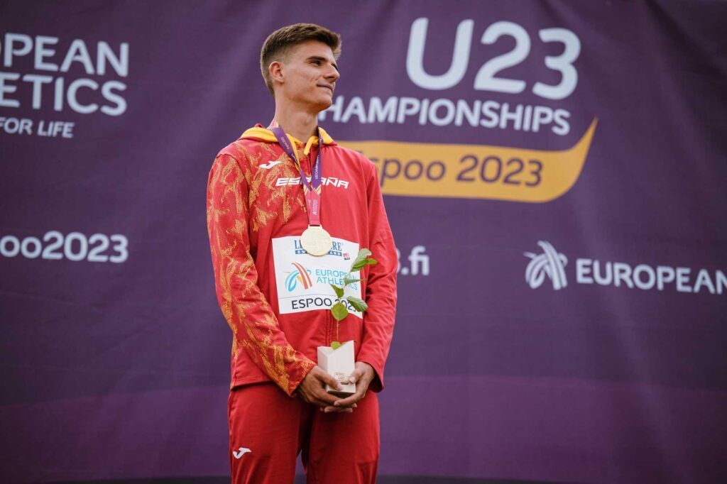 Un atleta, Alex Quijada, vestido con un chándal rojo de la selección española, posa en el podio con una medalla colgada al cuello y un pequeño trofeo en las manos, en el Campeonato Europeo de Atletismo Sub-23 celebrado en Espoo en 2023.