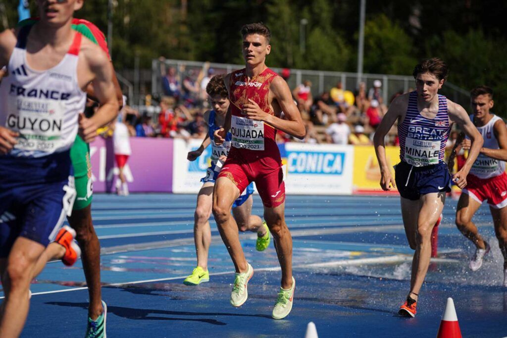 Los atletas corren por una pista azul durante una carrera, con Alex Quijada, vestido con un uniforme rojo, a la cabeza. En el fondo, iluminado por el sol, se ven espectadores y árboles, y se aprecian algunas salpicaduras de agua en la pista.