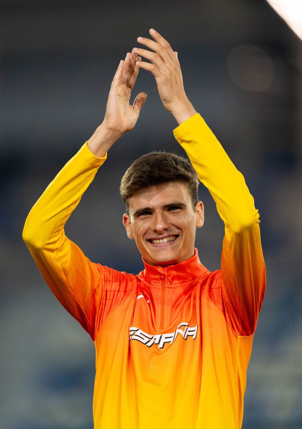 Alex Quijada, un deportista sonriente que lleva una chaqueta amarilla y naranja, aplaude por encima de la cabeza para celebrar. En la chaqueta se lee la palabra «España», lo que indica que representa a España. El fondo aparece desenfocado.